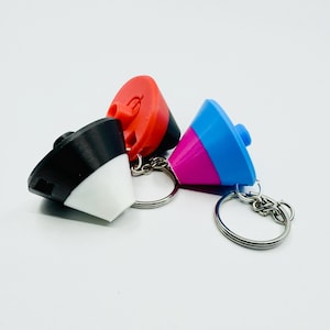 May include: Three colorful keychains with cone-shaped tops. One is black and white, another is red, and the third is blue, pink, and blue. Each keychain has a silver metal ring.