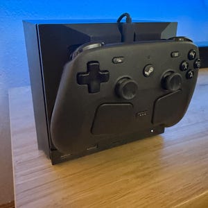 May include: A black video game controller with a central Steam logo button, a directional pad, two analog sticks, and four lettered action buttons, docked into a black rectangular charging station with a visible power cable connected at the top.