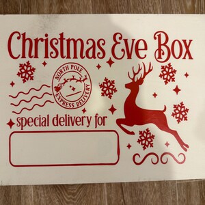 May include: A white wooden Christmas Eve Box with red lettering and graphics. The text includes "Christmas Eve Box", "North Pole Express Delivery", and "special delivery for". A reindeer, snowflakes, and stars decorate the box.