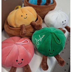 May include: A collection of plush dumpling toys with smiling faces and brown limbs. The set includes blue, yellow, white, pink, and green dumplings, with some nestled in brown baskets. The green dumpling features the white embroidered name Joshua.