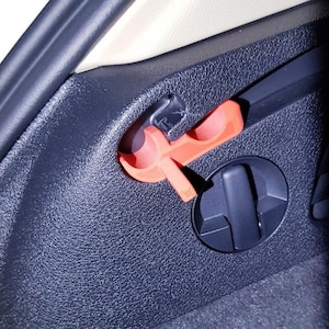 Peut inclure: Gros plan sur l'intérieur d'une voiture avec un panneau noir texturé et un outil en plastique orange vif inséré. L'outil a une forme unique avec un crochet et un levier. Un cadran circulaire noir est également visible.