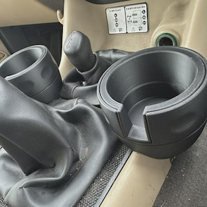 May include: Black 3D-printed cup holders designed for a vehicle's center console. The cup holders are cylindrical with a stepped design and a cutout for a handle. The surrounding area includes a gear shift and other vehicle components.