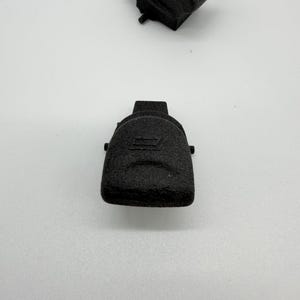 Könnte beinhalten: Zwei schwarze, strukturierte Kunststoffkomponenten, möglicherweise elektrische Anschlüsse, werden vor einem weißen Hintergrund gezeigt. Eine Komponente befindet sich oben, eine andere unten. Die Komponenten haben eine abgerundete, rechteckige Form mit strukturierter Oberfläche.