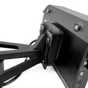 Puede incluir: Primer plano de un dispositivo electrónico negro montado en un soporte negro. El dispositivo tiene forma rectangular con esquinas redondeadas y varios botones y puertos pequeños. El soporte está fijado a una estructura de soporte más grande e inclinada.