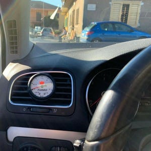 May include: A close-up of a car's dashboard featuring a turbo gauge. The gauge is round with a white face, black numbers, and a red needle. The word "TURBO" is printed on the gauge. The gauge is mounted in the air vent.