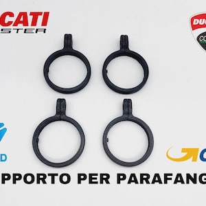 Könnte beinhalten: Vier schwarze Motorrad-Kotflügelhalterungen mit kreisförmigem Design und einer Befestigungslasche. Das Ducati Monster-Logo und der Text "SUPPORTO PER PARAFANGHI" sind ebenfalls sichtbar.