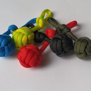 Può includere: Una collezione di portachiavi con nodi in paracord colorati. I portachiavi sono in vari colori, tra cui blu, giallo, rosso, nero e verde. Ogni portachiavi presenta un design a sfera annodata con un anello per l'attacco.