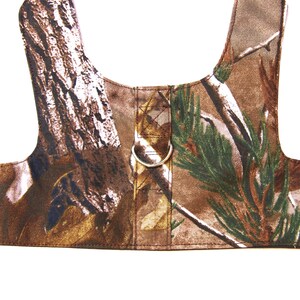 May include: Camouflage dog harness with a metal D-ring. The harness is made of brown, green, and white fabric with a woodland pattern.