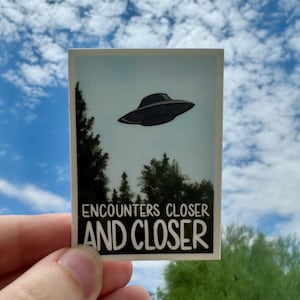 May include: A sticker with a black and white illustration of a UFO flying over a forest. The text on the sticker reads "Encounters Closer And Closer".