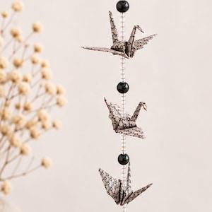 May include: A vertical hanging mobile featuring five folded paper origami cranes with a black and white patterned design. The cranes are strung on a clear cord, separated by alternating black spherical beads and clear beads, ending with a gold circular charm.