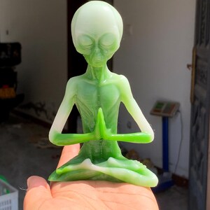 Puede incluir: Una figura de alienígena verde en una postura de yoga sentado con las manos juntas en oración. El alienígena tiene una cabeza grande, un cuerpo delgado y un degradado de verde y blanco. La figura se sostiene en una mano.