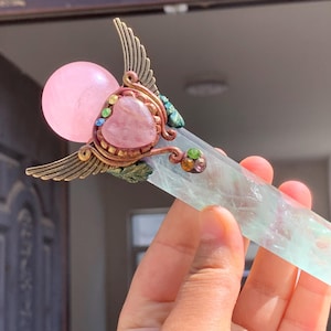Könnte beinhalten: Ein handgefertigter Kristallstab mit einer rosa Kugel, einem herzförmigen rosa Stein und einer klaren, mehrfarbigen Kristallspitze. Der Stab hat bronzefarbene Metallakzente mit flügelartigen Details und farbenfrohen Edelsteinverzierungen.