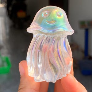 Puede incluir: Una figura de medusa clara e iridiscente con una cara sonriente. La figura tiene una parte superior redondeada y una parte inferior ondulada y fluida. Los colores cambian de azul a verde y rosa, creando un efecto brillante. La figura se sostiene en una mano.