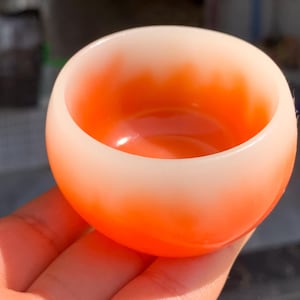 Puede incluir: Un cuenco pequeño y redondo con un degradado de color. El cuenco pasa de un borde blanco a un interior naranja vibrante. La superficie lisa y brillante refleja la luz, resaltando el contraste de color. El cuenco se sostiene en una mano.