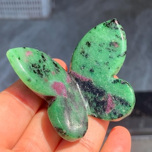 May include: A butterfly-shaped ornament carved from green stone, featuring black and red markings. The polished surface reflects light, and the wings are spread. The figurine is held in a hand, showcasing its detailed form.