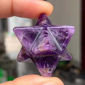 Può includere: Una stella Merkaba di ametista sfaccettata, tenuta tra le dita. Il cristallo è di un viola intenso con striature più chiare e motivi interni. La forma geometrica ha sei punte, ciascuna formata da facce triangolari. Oggetto spirituale o decorativo.