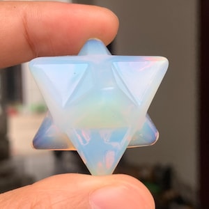 Op de afbeelding: Een doorschijnende, opalescente Merkaba-ster, een kristalformatie met een zespuntige stervorm. Het kristal heeft een zachte, iriserende gloed met hints van blauw en geel. De Merkaba wordt tussen de vingers gehouden.