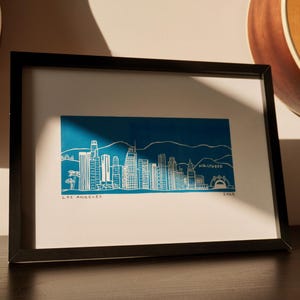May include: Framed art print featuring a white line drawing of the Los Angeles skyline against a blue background. The print includes the text "LOS ANGELES" and "HOLLYWOOD". The artwork is in a black frame.