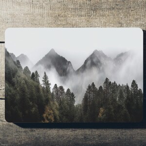 May include: A misty mountain landscape with a forest in the foreground. The mountains are covered in fog and the trees are dark green.