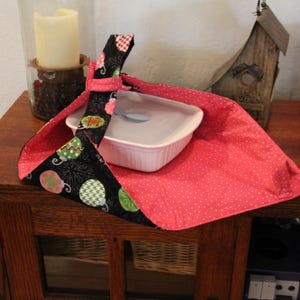 Puede incluir: Un portaplatos de tela con un diseño estampado en negro y rosa. El portaplatos está envuelto alrededor de una fuente rectangular blanca con tapa. El interior del portaplatos es de lunares rojos.