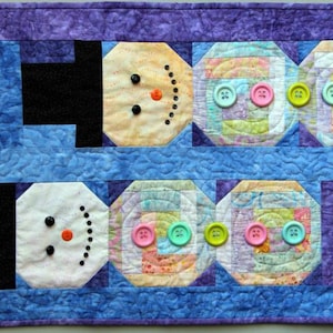 May include: A quilted wall hanging featuring two snowmen made of fabric scraps. The snowmen are layered with colorful buttons and have black top hats. The background is a purple and blue patchwork design. The text "Surrounded by Geese Over the Fields - QBN038" is at the bottom of the image.