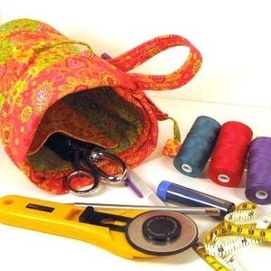 May include: A colorful sewing kit with a patterned fabric bag, scissors, spools of thread in blue, red, and purple, a yellow rotary cutter, and a measuring tape. The bag is open, revealing the contents.