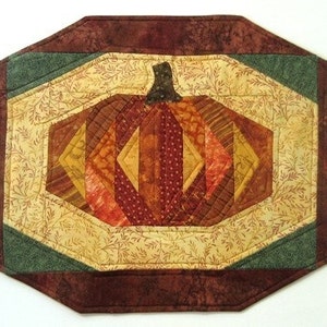 May include: A quilted table runner with a brown, green, and beige color scheme. The center of the runner features a patchwork pumpkin design.
