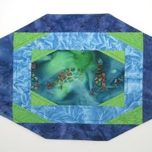 May include: A quilted table runner with a geometric design. The runner is made of blue, green, and white fabric. The center of the runner features a green and blue fabric with a brown and orange pattern.