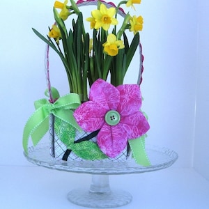 May include: A small, white and green patterned fabric basket with a pink fabric flower on top. The basket is filled with yellow daffodils and green leaves. The basket is sitting on a clear glass cake stand.