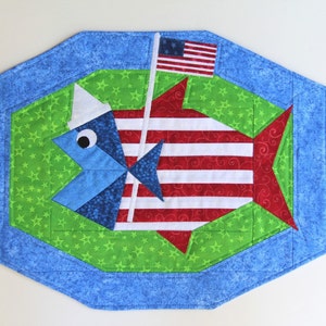 May include: A red, white, and blue fish-shaped table runner with a green background and a small American flag on a stick. The fish is wearing a blue and white sailor hat.