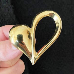 Puede incluir: Un broche dorado en forma de corazón. El broche tiene una superficie pulida y reflectante y un diseño curvo y abierto. El corazón se sostiene sobre un fondo oscuro.
