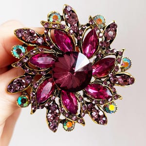 Puede incluir: Broche con forma floral con una gran piedra central facetada de color púrpura oscuro. El broche presenta piedras en forma de pétalo en varios tonos de púrpura y piedras de acento iridiscentes más pequeñas, todo en un marco de metal dorado.