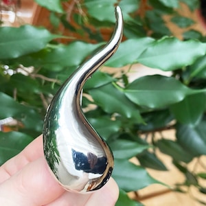 May include: A polished, teardrop-shaped metal sculpture with a curved, elongated top. The sculpture has a reflective, silver finish, and the smooth surface reflects the surrounding greenery. The design is abstract and modern.