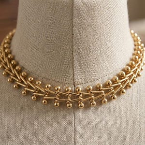 Może przedstawiać: Złoty naszyjnik typu choker z powtarzającym się geometrycznym wzorem połączonych metalowych pręcików i małych kulistych koralików. Konstrukcja składa się z szeregu połączonych segmentów, które tworzą teksturowany wygląd przypominający łańcuch.