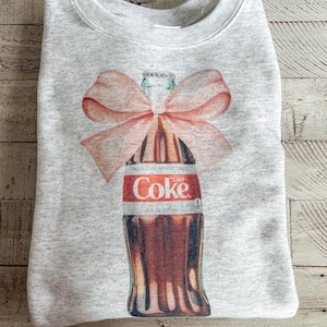 Könnte beinhalten: Ein graues Sweatshirt mit einem Aufdruck einer Glasflasche Diet Coke, die mit einer rosafarbenen Schleife gebunden ist. Die Flasche trägt den Schriftzug "Diet Coke" auf dem Etikett.