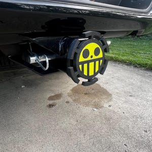 Peut inclure: Un cache-attelage noir avec un motif de crâne de dessin animé jaune et noir. Le cache est fixé à un véhicule noir. Le motif présente un visage jaune avec des yeux et une bouche noirs, entouré d'une bordure noire.