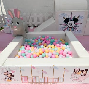 May include: A white ball pit filled with pastel-colored balls. The pit features a design with Mickey and Minnie Mouse, the text "Leilaland", and the phrase "The Happiest Place On Earth". A gray bunny toy sits nearby.