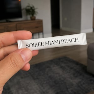 Può includere: Una bustina bianca rettangolare a forma di bastoncino con bordi frastagliati su entrambe le estremità. Il testo SOIRÉE MIAMI BEACH è stampato con un carattere serif nero al centro della superficie bianca. La bustina è tenuta orizzontalmente davanti a uno sfondo sfocato.