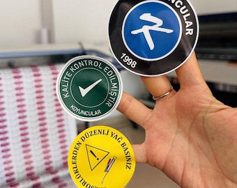 May include: A hand holding three round stickers. The top sticker is black with a blue circle and white text and a symbol. The middle sticker is green with a white checkmark. The bottom sticker is yellow with a warning symbol.