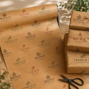 Puede incluir: Un rollo de papel de embalaje kraft marrón con un patrón repetitivo de logotipos de Mono Coffee Roasters, Luna Bakery y Verde & Co. Packaging Solutions. El papel se muestra en un dispensador de metal con tres paquetes envueltos con cordel.