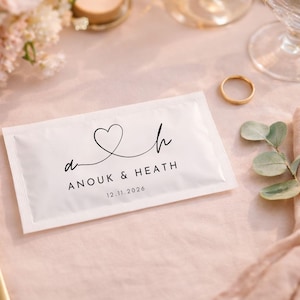 May include: A rectangular white packet featuring black printed text that reads ANOUK & HEATH, the date 12.11.2026, and a stylized line drawing of a heart connecting the lowercase letters a and h. The packet is centered on a light pink fabric surface.