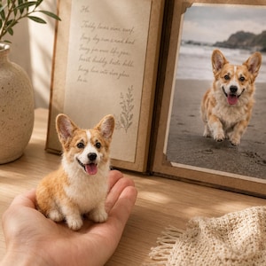 Puede incluir: Una pequeña figura de Corgi galés de Pembroke hecha de lana fieltrada, con pelaje color canela y blanco, sentada. A su lado hay un álbum de fotos abierto de estilo rústico con una nota manuscrita a la izquierda y una foto de un Corgi.