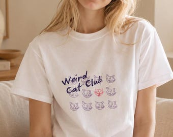 May include: A white short-sleeved t-shirt featuring the text "Weird Cat Club" in dark blue script, accompanied by a grid of twelve small, simple cat face illustrations in blue, with one cat face highlighted in red ink.