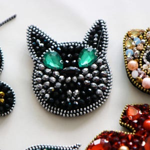May include: A black cat face brooch with green gem eyes and silver beads. The brooch is made of fabric and beads.