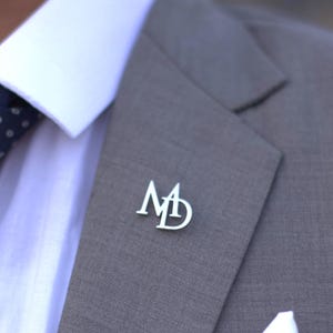 May include: A silver lapel pin with the intertwined letters "MD" is affixed to a gray suit jacket. The jacket is paired with a white dress shirt and a navy blue tie with white polka dots. A white pocket square is visible.