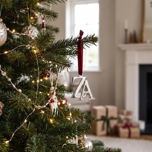 Puede incluir: Un adorno de metal plateado con las letras Z y A en una fuente con serifa, suspendido por una cinta de terciopelo rojo oscuro. El adorno cuelga de una rama de pino en un árbol de Navidad decorado con luces blancas cálidas.