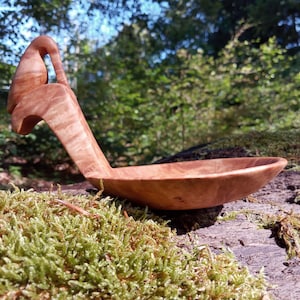Op de afbeelding: Een handgesneden houten lepel met een gladde, lichtbruine afwerking en zichtbare natuurlijke houtnerven. Het handvat is gevormd tot een gestileerde vogelvorm die omhoog steekt vanuit de ondiepe, ovaalvormige kom van de lepel.