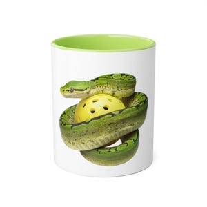 Può includere: Tazza in ceramica bianca con interno verde. Un serpente verde con segni neri è avvolto attorno a una palla gialla. Un oggetto regalo originale.
