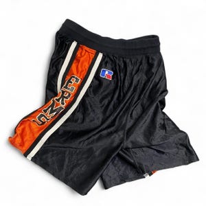 Puede incluir: Pantalones cortos deportivos negros con cintura elástica, un panel lateral en naranja brillante con ribete blanco y la palabra ERMU impresa verticalmente en negro. Un pequeño parche con el logo de la marca Russell en rojo, blanco y azul.