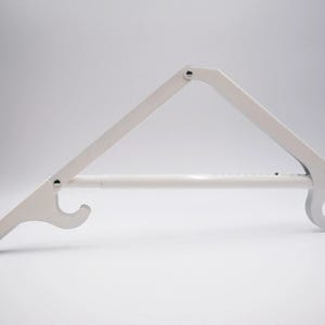 May include: A white metal folding shelf bracket featuring a triangular support arm and a horizontal bar. The bracket includes two curved hook-shaped ends for mounting and is constructed with visible metal rivets at the pivot points.